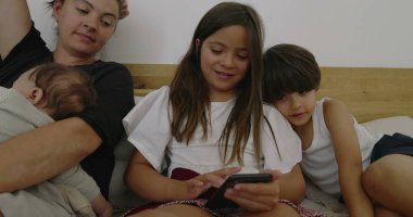 Anne kucağında uyuyan bebek, kız ve oğul akıllı telefona odaklanmış, aile yatakta bir dakika paylaşıyor, rahat bir yuva kuruyor, kardeş etkileşimi, modern aile dinamikleri