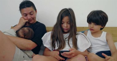 Bir anne, büyük çocukları telefona odaklanırken bebeğini emziriyor, yatakta samimi bir aile anı yaratıyor, evde bir birliktelik ve rahatlama hissi yaratıyor..