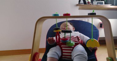 Bebek bir fedainin içinde uzanıyor ve asılı oyuncaklarla oynuyor, rahat bir ev ortamında keşiften ve oynamaktan zevk alıyor, çocukluk yıllarının merak ve masumiyetini vurguluyor..