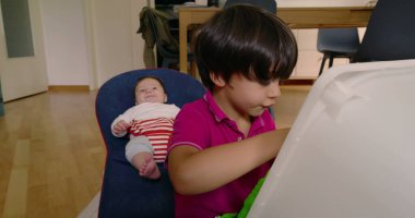 Büyük kardeş oyuncakla oynarken küçük kardeş bir fedainin içinde dinleniyor, aile hayatının bir anını yakalıyor ve rahat bir ev ortamında kardeş etkileşimi kuruyor..