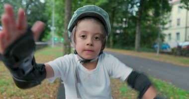 Kaykayın üzerinde koruyucu kıyafetlerle oturan bir çocuk, kaskını takmış, sakin bir ifadeyle kameraya bakıyor, park SD 480p 'sinde ağaçlar ve yeşilliklerle çevrili.