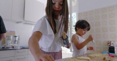 Çocuklar mutfakta hamur hazırlıyor, kız odaklanıyor, oğlan asistanı, aile yemekleri aktivitesi, anne arka planda, parlak ev dekorasyonu, etkileşimli öğrenme anı, takım çalışması 480p