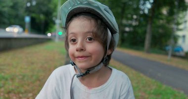 Koruyucu teçhizatlı bir çocuk yol kenarında kaykayın üzerinde oturuyor, kaskını takıyor, geçen arabalara bakıyor, yeşil ve ağaçlarla çevrili, bulutlu bir günde.