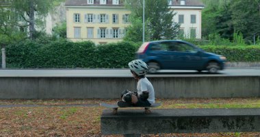 Koruyucu teçhizatlı bir çocuk yol kenarında kaykayın üzerinde oturuyor, kaskını takıyor, geçen arabalara bakıyor, yeşil ve ağaçlarla çevrili, bulutlu bir günde.