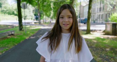 Uzun saçlı bir kız ileri doğru yürüyor ve sonra geri dönüp kameraya bakarken ağaçlarla kaplı patikaları ve arka planda binaları olan güneşli bir parkta yürüyor.