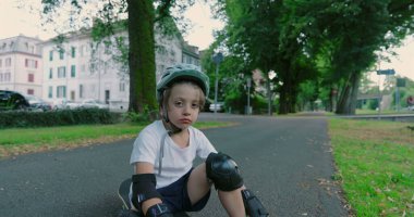 Kaykayın üzerinde koruyucu giysilerle oturan, kask takan bir çocuk, bulutlu bir günde çevrenin yeşillik ve binalarla çevrili olduğu bir yerleşim alanındaki ağaçlık yolda ciddi görünüyor.