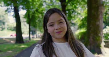 Parkta gülümseyen, beyaz bir bluz giyen, arka planda bulanık yeşillik ve ağaçlarla dolu uzun saçlı genç bir kızın yakın çekimi mutlu ve huzurlu bir anı yakalıyor.