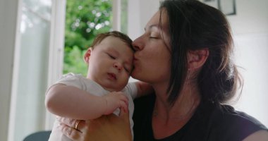 Anne bebeğini kollarında tutarken yanağından öpüyor, şefkatli ve sevgi dolu bir an yaratıyor, sıcak bir ev ortamında güçlü bir bağ ve sevgiyi vurguluyor - SD 480p