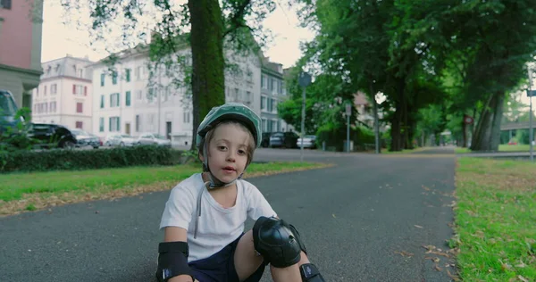 Kaykayın üzerinde koruyucu giysilerle oturan, kask takan bir çocuk, bulutlu bir günde çevrenin yeşillik ve binalarla çevrili olduğu bir yerleşim alanındaki ağaçlık yolda ciddi görünüyor.