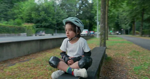 Koruyucu teçhizatlı bir çocuk yol kenarında kaykayın üzerinde oturuyor, kaskını takıyor, geçen arabalara bakıyor, yeşil ve ağaçlarla çevrili, bulutlu bir günde.