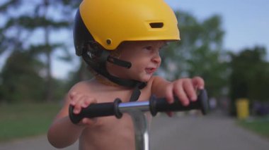 Motosikletli çocuk, sarı kasklı, gidonu tutan ve parktaki çevreyi izleyen meraklı, gözlemci ve erken motor beceri geliştiren biri..