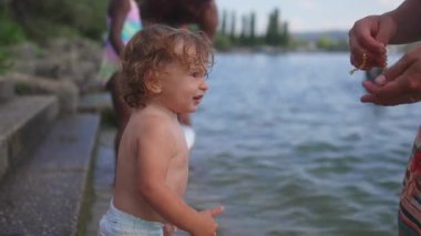 Göl kenarında dikilmiş, annesinin bileğini işaret eden çocuk, merak ve bileklikle oynama arzusunu gösteriyor, sakin bir yaz ortamında çevreyi keşfederken..