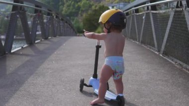 Yürüyen çocuk, yaya köprüsünde sarı kaskla scooter kullanıyor. Dengeyi, bağımsızlığı ve merakı açık hava yaz aktiviteleri sırasında etrafı keşfederken gösteriyor..