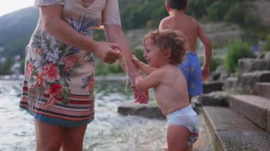 Gölün kenarında duran küçük çocuk, anne bileziğine uzanırken, büyük kardeş yakınlarda duruyor. Açık hava ortamında merak, etkileşim ve aile bağlarını gösteriyor..