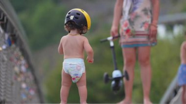 Köprüde sarı başlık ve bez takmış küçük bir çocuk scooter 'ın yanında duruyor. Bu arada anne de deniz kenarında neşeli bir yaz ailesi anı izliyor..
