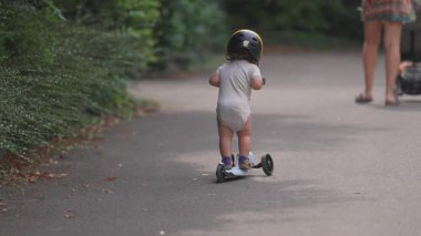 Motosikletli bir çocuk, annesine bakmak için geri dönüyor. Bebek arabası itiyor. Park yolunda neşeli bir aile anı..