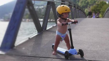 Küçük çocuk, sarı başlık takarak scooter 'ın üzerinde duruyor, küçük çocuk köprü rampasının başında duruyor, odaklanarak ileriye bakıyor, dışarıda neşeli bir yaz öğleden sonrasında eğleniyor..