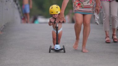 Anne, çocuğun scooter 'da durmasına yardım ediyor, sarı kasklı küçük bir çocuk yaya köprüsünde, eğlenceli açık hava yaz aile anı sırasında yavaşlıyor.