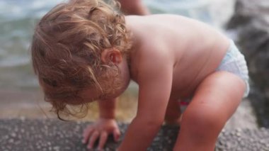 Su kenarında taş yüzeyini keşfeden küçük çocuk, denge için öne doğru eğiliyor, merak, keşif ve erken duyusal gelişimi gösteriyor.