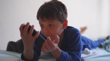Genç çocuk elinde akıllı telefonla yatakta uzanıyor. İçeriği izlerken ya da oyun oynarken odaklanmış ifade ediyor. Evde sakin ve sakin bir ekran zamanı.