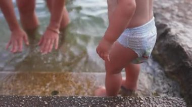 Göl kenarındaki taş basamaklarda duran bebeğe yakın çekim yaparken, anne destek için ellerini suya uzatarak, güvenliği, dengeyi ve ebeveyn korumasını vurguluyor.