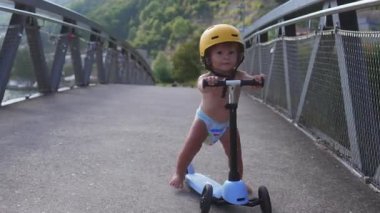 Küçük çocuk köprüden aşağı skuter sürerken sarı kask takıyor. Küçük çocuk dışarıda dengede duruyor. Eğlenceli yaz aile anlarını keşfediyor.