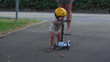 Banliyö yolunda sarı kasklı küçük bir scooter süren küçük çocuk, neşeli bebek öğrenme dengesi anne tarafından denetlenirken, yaz parkında erken çocukluk aktivitesi