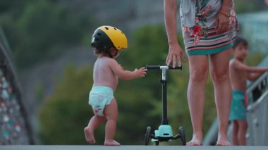 Yürüyen çocuk köprüde annesinin yanında scooter sürüyor. Sarı kasklı küçük çocuk dışarıda dengeyi kurmayı öğreniyor. Yazın eğlenceli aile anlarını keşfediyor.