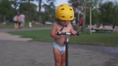 Parkta scooter süren küçük bir çocuk, sarı başlık takıyor, kaldırımları keşfeden küçük bir çocuk, neşeli bir merakla, güneşli yaz öğleden sonranın tadını çıkarıyor.