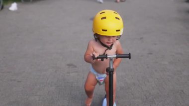 Sarı kask takan, elinde gidonu tutarken durmak ve geriye bakmak için duraklayan, açık hava yaz etkinliği sırasında merak ve farkındalık gösteren bir çocuk.