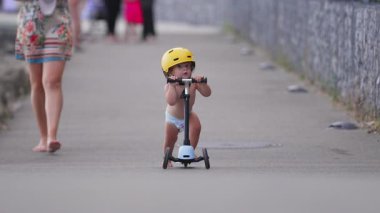 Anne arkada yürürken yolda sarı kaskla scooter süren çocuk, erken bağımsızlık, merak ve açık havada çocukluk keşiflerini gösteriyor.