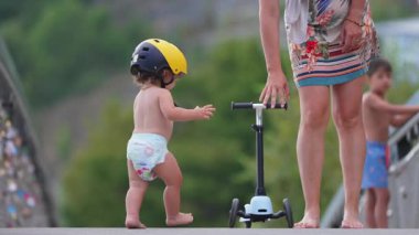 Anne motosikleti yerleştirir, bebek sarı kaskla yokuş aşağı gider, anne onu durdurmaya çalışır ama o düşer ve ağlamaya başlar.