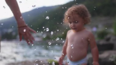 Su sıçrarken göl kenarındaki çevreyi gözleyen küçük çocuk, sakin bir açık hava yaz ortamında merak, keşif ve erken çocukluk merakını gösteriyor.
