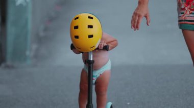 Küçük çocuğun sarı kask takıp, scooter sürmeyi öğrenirken, anne gidonu frenlerken, koruma, güvenlik, bağ ve erken çocukluk keşiflerini ön plana çıkarırken.