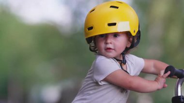 Yürüyen çocuk scooter 'ı geride bırakıyor ve sarı başlık takarak ileri doğru yürüyor. Küçük çocuk açık havada eğlenceli bir yaz öğleden sonrası boyunca merak ile park yolunu keşfediyor.