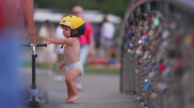Küçük çocuk, sarı başlık takarak scooter 'a doğru yürüyor. Küçük çocuk, açık havada eğlenceli yaz aile anları sırasında renkli aşk kilitleriyle köprüyü keşfederken annesine uzanıyor.