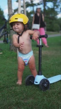 Sarı miğfer takan küçük bir çocuk parktaki yeşil çimlerde scooterla duruyor, mutlu ve meraklı ifade, açık hava eğlence aktivitesi, erken çocukluk gelişimi, yaz oyun zamanı