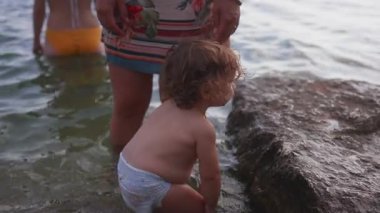 Büyük kayaların yanında sığ sularda ayakta duran küçük çocuk. Anne ise yanında duruyor. Ebeveynlerin rehberliğini, güvenliğini ve göl kenarındaki açık havada yapılan çocukluk keşiflerini gösteriyor.