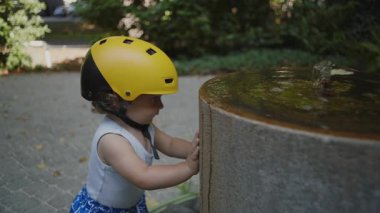 Dışarıdaki taştan fıskiyeye dokunan çocuk sarı miğfer takıyor, kaynayan suyu merakla izliyor, yazın neşeli keşifleri ve keşiflerinin ilk zamanlarını izliyor.
