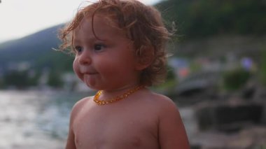 Gölün kenarındaki kıvırcık saçlı çocuğun yakın çekimi, yumuşak akşam ışığıyla düşünceli bakışları, açık havada huzurlu bir çocukluk anı.