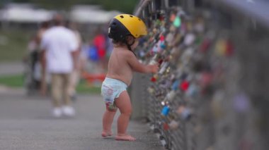 Sarı miğfer takan küçük çocuk kilitlerle dolu çitlerin yanında duruyor, dışarıdaki eğlenceli yaz aktiviteleri sırasında merak ve hayranlıkla çevreyi keşfediyor.