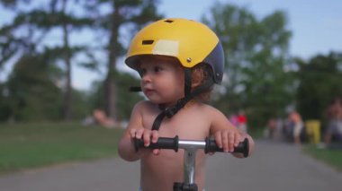Motosikletli bir çocuk sarı bir miğfer takıyor, meraklı bir şekilde yan tarafa bakıyor, dışarıdaki park yollarını keşfederken çevreyi inceliyor yaz etkinlikleri sırasında.