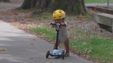 Odaklanmış ifade ile scooter süren küçük çocuk, sarı kasklı küçük çocuk yaz boyunca park yolunda denge ve koordinasyon çalışması yapıyor ve dışarıda oyun oynuyor.