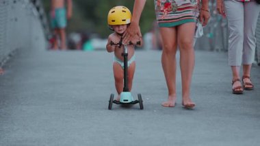 Annesi yanında yürüyüp rehberlik, güvenlik ve erken çocukluk motor gelişimini gösterirken, mobiletinde sarı başlıklı bir çocuk gidonu tutuyordu.