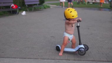 Küçük çocuk skuterin üzerinde sarı başlık takıyor, küçük çocuk kaldırım yolunda duruyor gidonu tutuyor, dışarıda neşeli bir yaz öğleden sonrasında eğleniyor.