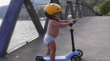 Scooter 'ın üzerinde sarı kaskla duran, gidonu tutan ve köprünün üzerinde durup çevreyi izleyen, merakını ve erken motor becerilerini gösteren bir çocuk.