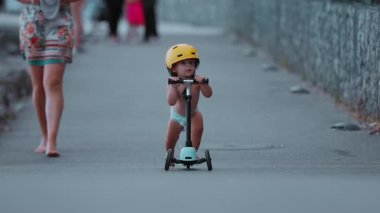 Küçük çocuk park yolunda skuter sürerken sarı miğfer takıyor, küçük çocuk kendinden emin bir şekilde öne doğru sürüyordu.