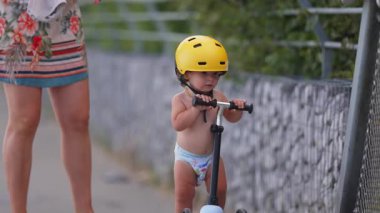 Motosikletli bir çocuğun yakın çekimi sarı başlık takarken annesi gidonu nazikçe fren yapar, güvenlik, koruma ve erken çocukluk eğitimine odaklanır.