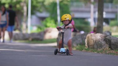 Küçük çocuk park yolunda skuter sürerken sarı kask takıyor, küçük çocuk açık havada yaz aktivitelerinin keyfini çıkarırken denge ve koordinasyon çalışıyor, oyuncu yaşam tarzı anı.