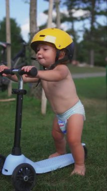 Parkta çimenlerde scooter süren küçük çocuk sarı kask takıyor, odaklanmış ve kararlı ifade takınıyor, açık havada oyun zamanı, erken çocukluk hareketliliği ve denge gelişimi